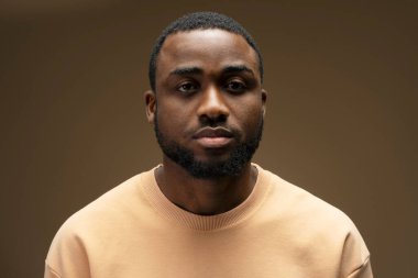 Kendine güvenen Afro-Amerikan erkek modelin günlük giysiler giydiği bir stüdyo portresi. Kahverengi arka plana karşı oluşturduğu yüz ifadesiyle ciddiyetini ve kararlılığını ifade ediyor.