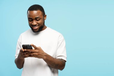 Beyaz tişörtlü mutlu Afro-Amerikan bir adam interneti gezmek, sohbet etmek, mesajları okumak ve mavi bir stüdyoda oyun oynamak için cep telefonu kullanıyor. Çevrimiçi teknoloji kavramı