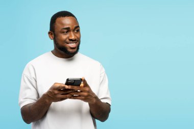 Beyaz tişörtlü genç Afrikalı bir adam, cep telefonu kullanırken gülümsüyor ve mavi bir arka plana bakıyor, gelişigüzel bir ortamda neşe ve bağlantı yaratıyor.