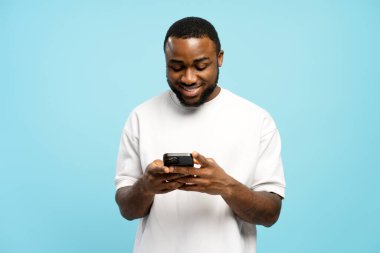 Beyaz tişört giyen, akıllı telefon kullanan ve canlı mavi bir stüdyo geçmişine karşı gülümseyen mutlu genç Afrikalı Amerikalı adam. Çevrimiçi alışveriş kavramı