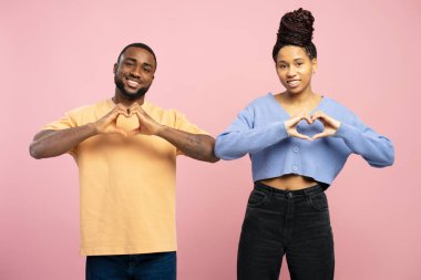 Gülümseyen Afro-Amerikan erkek ve kadın ellerinde sevgi ve özen göstererek kalp hareketi yapıyorlar pembe stüdyo arka planında duruyorlar samimi duygular gösteriyorlar, yürekten duygular gösteriyorlar.