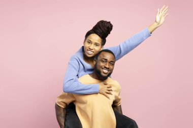 Neşeli Afrikalı Amerikalı adam, güzel kız arkadaşına sırtına biniyor. O da gülümseyen ve pembe arka plandaki kameraya bakarken birine el sallıyor. Eğlenceli konsept