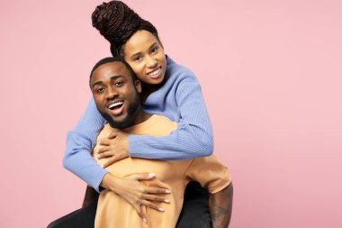 Neşeli genç siyahi kadın, onu sırtında taşırken, erkek arkadaşına neşeyle sarılıyor. İkisi de canlı pembe stüdyo geçmişine gülümsüyor. Mutluluk ve sevgi saçıyor.