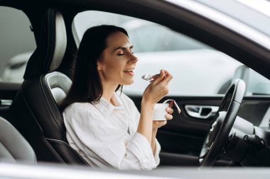 İş kadını arabasında rahat bir şekilde oturup yoğun bir programa ara verirken lezzetli bir yoğurdun tadını çıkarıyor ve sağlıklı beslenmenin tadını çıkarıyor.