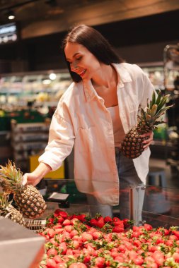 Güzel, gülümseyen bir kadın, marketin meyve reyonundan taze ananas ve çilek seçen bir müşteri sağlıklı malzemeler alırken gülümsüyor.