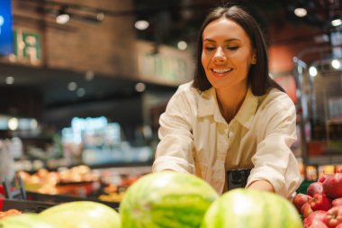 Genç bir kadın bir marketin meyve ve sebze reyonundan olgun bir karpuz seçerek sağlıklı gıda alışverişi ve taze ürün deneyiminin tadını çıkarıyor.