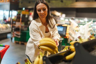 Mutlu müşteri, Latin kadın marketteki bir vitrinden taze muz toplarken gülümsüyor besleyici bir diyet için sağlıklı ve olgun meyveler seçme deneyiminin tadını çıkarıyor.