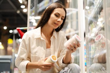 Latin bir kadın iki yoğurt kavanozunu karşılaştırıyor, marketin dondurulmuş süt ürünleri bölümünde karar veriyor, dikkatli alışveriş alışkanlıkları gösteriyor. Alışveriş konsepti