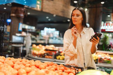 Akıllı telefon kullanan ve süpermarkette mandalina seçen, market alışverişi yapan ve alışveriş listesini kontrol etmek için mobil uygulama kullanan genç bir kadın. Gıda, çevrimiçi teknoloji kavramı