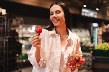 Mutlu müşteri gülümsüyor ve marketten taze çilek seçiyor, bir sepet çilek tutuyor ve aldığı çileğe bakıyor, süpermarketten sağlıklı yiyecekler alıyor. Yiyecek konsepti