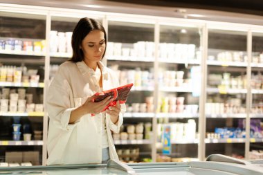 Çekici Latin kadın, modern bir süpermarketin soğutucu koridorunda alışveriş yaparken dondurulmuş gıda paketinin etiketini dikkatle okuyor ve bilgilendirilmiş satın alma kararları veriyor.