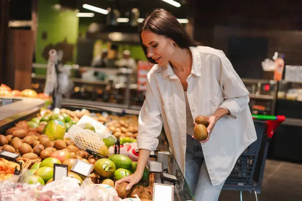 Genç bir kadın, modern bir süpermarketin üretim bölümünden taze kivi meyvesi seçiyor, bir elinde kivi tutarken diğer elinde daha fazla meyve oluyor.