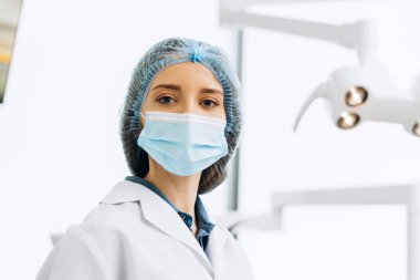 Diş muayenehanesinde poz verip kameraya bakan koruyucu şapkalı ve maskeli neşeli bir doktorun portresi. Diş sağlığı, sağlık hizmetleri konsepti