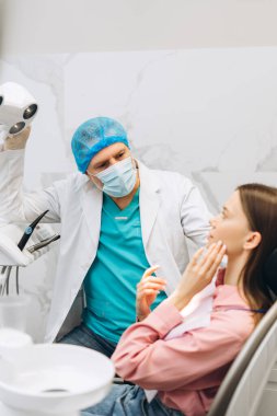 Erkek dişçinin portresi steril maske takıyor dişçi lambasını dişçi muayenehanesinde kadın hasta için kullanıyor. Diş sağlığı, sağlık hizmetleri konsepti