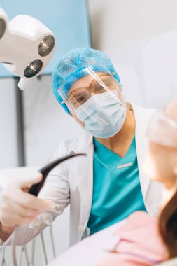Klinikteki kadın hastaya diş tedavisi sırasında diş aletleri kullanan yüz maskeli erkek dişçinin portresi. Sağlık hizmetleri diş tedavisi