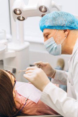 Yüz maskesi takan ve eldiven takan olgun dişçinin portresi. Kadın dişlerini aynayla kontrol ediyor ve dişçi kliniğine giriyor. Diş sağlığı, sağlık hizmetleri konsepti