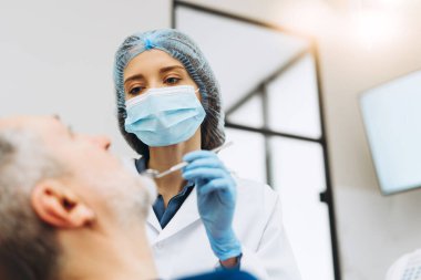 Koruyucu yüz maskesi takmış kadın doktor portresi modern diş kliniğinde aynayla erkek dişlerini kontrol ediyor. Sağlık hizmetleri diş tedavisi