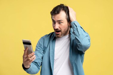 Akıllı telefonuna bakarken kafasını tutan şaşırmış bir adam, beklenmedik bir habere, parlak sarı bir arka planda şok edici bir ifadeyle tepki veriyor. Çevrimiçi teknoloji kavramı