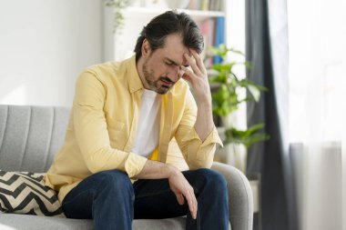 Olgun bir adam evdeki kanepede oturuyor, depresyonda ve yalnızlık çekiyor, elleriyle alnına dokunuyor, zihinsel sağlık sorunlarıyla başa çıkıyor.