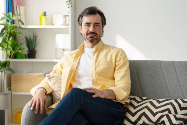 Rahat orta yaşlı bir adam şık, modern bir oturma odasındaki gri bir koltukta rahat bir şekilde oturur, ev ortamından memnuniyet ve güven yayar. Reklam kavramı