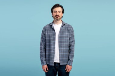 Sakallı, kareli gömlek giyen orta yaşlı bir adam, mavi bir arka plana karşı poz veriyor, rahat ve şık bir şekilde gündelik tarz ve olgun bir erkeklik örneği.