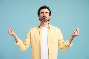 Sarı gömlekli beyaz adam, meditasyon yaparken gözlerini kapadı ve ellerini kaldırdı. İç huzurunu ve farkındalığını canlı mavi arka planda buldu. Zihinsel sağlığı ve stres atılımını destekledi.