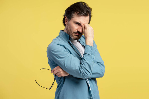 Стрессовый бизнесмен, страдающий от головной боли, массирующий мост носа, держащий очки в руке, стоящий на желтом фоне, одетый в синюю рубашку, имеющий болезненное выражение лица