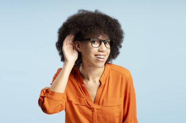 Gözlüklü, turuncu tişörtlü ve mavi arka planda eli kulağına dolanmış genç Afrikalı Amerikalı kadının stüdyo portresi.