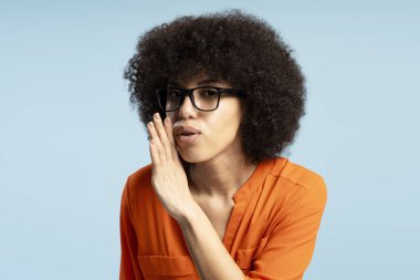 Genç Afrikalı Amerikalı iş kadını portresi canlı mavi bir arkaplan karşısında bir sır fısıldıyor, ilginç bir ifade ve ince el hareketleriyle dedikoduları paylaşıyor. Gizli konsept