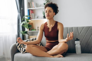 Huzurlu genç kadın kanepesinde yoga ve meditasyon yapıyor, rahat bir ev ortamında farkındalığı ve rahatlığı kucaklıyor, iç huzuru ve refahı geliştiriyor.