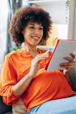 Mutlu hamile kadın kanepeye oturup bebek isimleri yazıyor, hamileliğin tadını çıkarıyor, anneliğe hazırlanıyor, geleceği planlıyor, modern bir apartman dairesi tasarlıyor. Çocuk doğumu kavramı