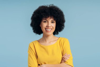 Sarı tişörtlü, kolları mavi arkaplanda çapraz duran neşeli ve kıvırcık saçlı Afrikalı Amerikalı bir kadının stüdyo portresi. Reklam kavramı