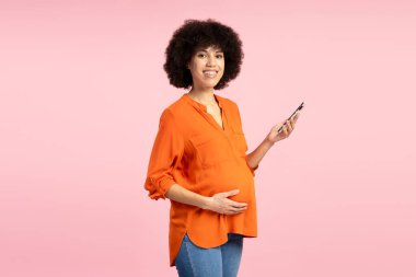 Mutlu gülümseyen Latin hamile kadın akıllı bir telefon tutuyor ve pembe arka planda neşeyle gülümserken nazikçe karnına dokunuyor, sevgi saçıyor. Annelik kavramı