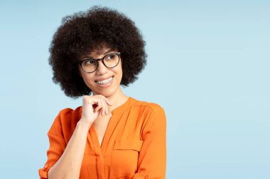 Turuncu gömlek, gözlük ve diş teli takan, çenesine eliyle dokunan ve mavi arka plana bakan düşünceli genç Afrikalı Amerikalı kadının stüdyo portresi. Reklam kavramı