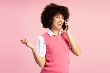Kıvırcık saçlı neşeli genç Latin kadın akıllı bir telefonla konuşuyor ve elini hareket ettiriyor. Canlı, pembe bir fon karşısında yapılan konuşmada heyecan ve nişanı dile getiriyor.