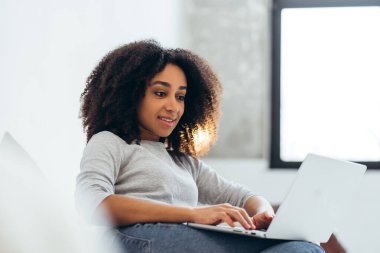 Uzaktan bilgisayarla çalışan gülümseyen bir kadın. Parlak oturma odasında rahat bir ofis kuruyor. Verimliliği ve rahatlığı kucaklıyor.