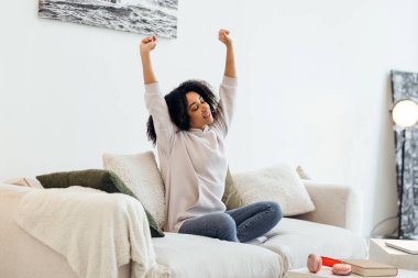 Mutlu Afrikalı Amerikalı kız rahat kanepesinde gözleri kapalı, kollarını kaldırmış, temiz hava soluyor, modern oturma odasında dinleniyor.