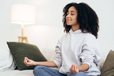 Huzurlu genç kadın meditasyondan zevk alıyor, rahat oturma odasındaki rahat koltukta iç huzuru ve dengeyi buluyor.