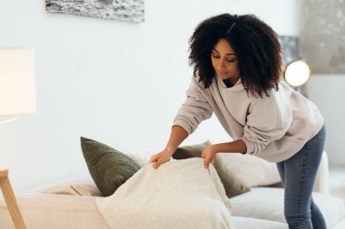 Beyaz tüylü battaniye ve yeşil yastıkları parlak oturma odasındaki bej koltuğa yerleştiren ev hanımı rahat ve davetkar bir ortam yaratıyor.