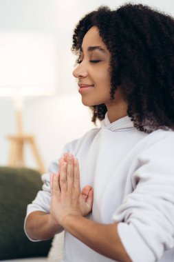 Huzurlu genç bir kadın, elleri ve gözleri kapalı meditasyon yapıyor, dua pozisyonunda birleşiyor, yoga pratiğiyle iç huzurunu ve farkındalığı buluyor.