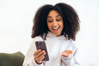 Heyecanlı genç kadın evde cep telefonu kullanıyor, mesajda ya da e-postada iyi haberler okuyor, online galibiyeti ya da indirimi kutluyor, şaşırmış ve mutlu hissediyor