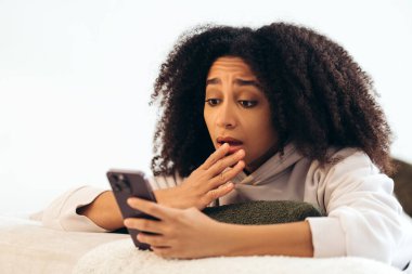 Kıvırcık saçlı genç bir kadın akıllı telefonunda beklenmedik bir mesaj okurken şok halinde inanılmaz bir habere tepki veriyor.