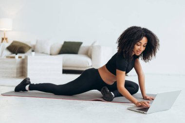 Çevrimiçi fitness kursu için dizüstü bilgisayar kullanırken evde esneme egzersizleri yapan sağlıklı ve sağlıklı bir kadın.