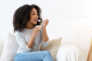 Gülümseyen Afrikalı Amerikalı bir kadın plastik bardaktan yoğurt yiyor ve evinde rahat bir koltukta oturuyor.