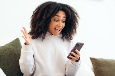 Heyecanlı genç bir kadın elinde akıllı telefonuyla online zaferi kutluyor, başarı ya da iyi haber, şaşırmış kız evde oturmuş cep telefonu uygulaması kullanıyor.