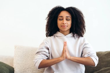 Huzurlu kadın kapalı gözlerle farkındalık yapıyor, evindeki rahat bir kanepede iç huzurunu buluyor.