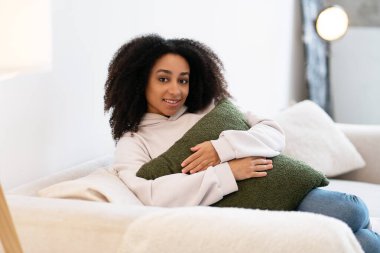 Huzurlu genç kadın rahat bir kanepede dinleniyor, oturma odasında yumuşak bir yastığı kucaklıyor.