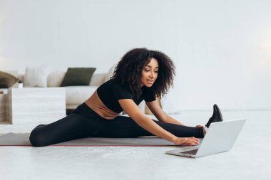 Sporcu kadın dizüstü bilgisayarındaki online talimatları takip ederek evde esnekliğini ve gücünü artırarak yan yana geriniyor.