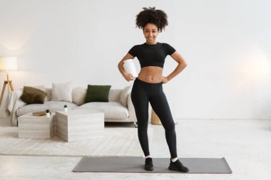 Sportif Afrikalı Amerikalı kadın besleyici protein takviyesi tutuyor, yoga minderinde poz veriyor, ev içi eğitime hazır.