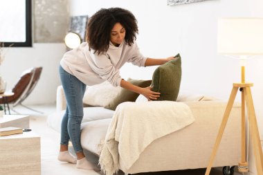 Kadın modern oturma odasını düzenliyor, rahat bir kanepeye yastıklar yerleştiriyor, rahat ve davetkar bir ortam yaratıyor.